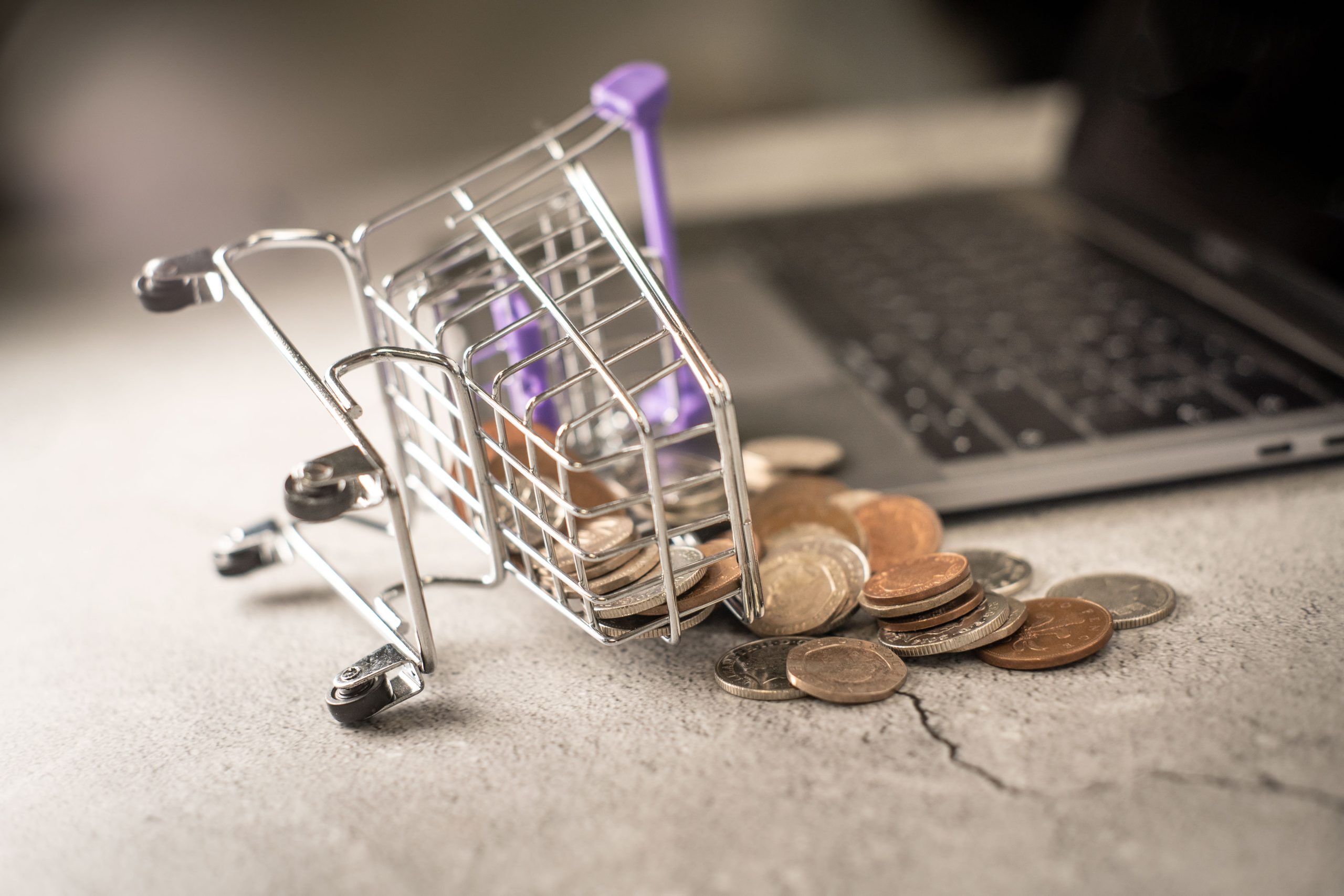 A tiny shopping cart with coins spilling out beside a laptop keyboard, representing the financial risk of fake online stores.