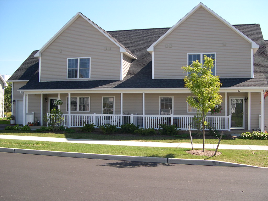 A beige two-story duplex with vinyl siding, a covered front porch, and a dark asphalt shingle roof on a residential street